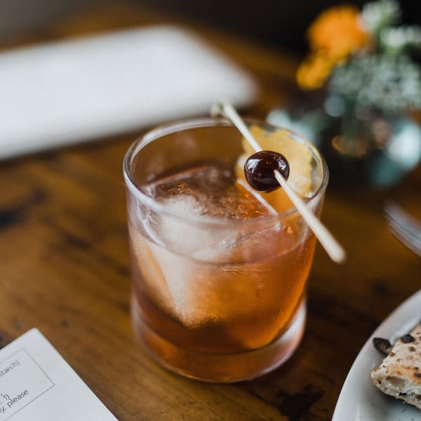 A cocktail with ice, garnished with an orange slice and cherry, next to a slice of pizza.