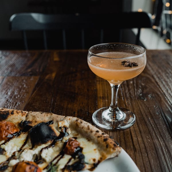 A wood-fired pizza with cherry tomatoes and mozzarella, next to a cocktail on a wooden table.