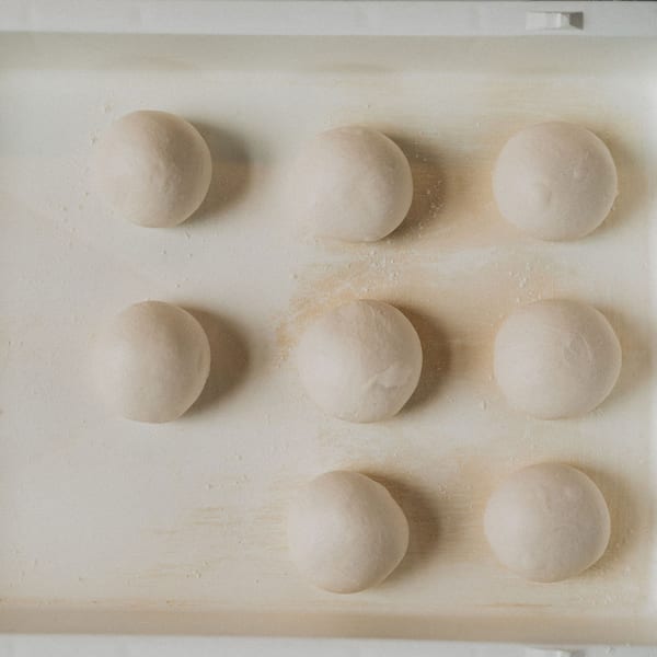 Nine balls of fresh pizza dough, lightly floured, resting on a light-colored surface.