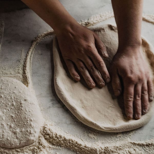 Hands stretching pizza dough on a floured marble surface, with another dough ball nearby.