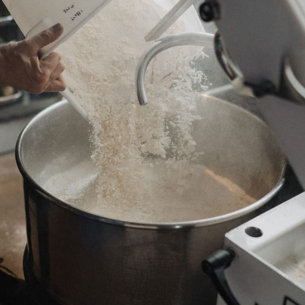 A hand pours flour into a large industrial mixer, preparing dough for Ovn Wood Fired Pizza.