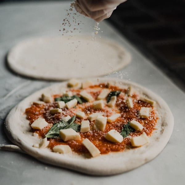 A hand sprinkles flour over an uncooked Ovn Wood Fired Pizza with sauce, cheese, and basil.