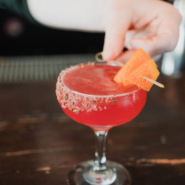 A bartender garnishes a vibrant red cocktail with an orange slice and a salt-rimmed glass.