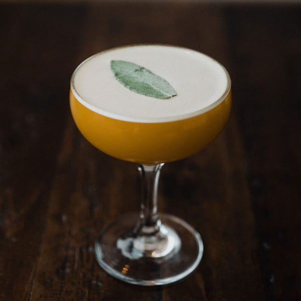A golden cocktail with a frothy top and a single sage leaf garnish on a dark wooden table.