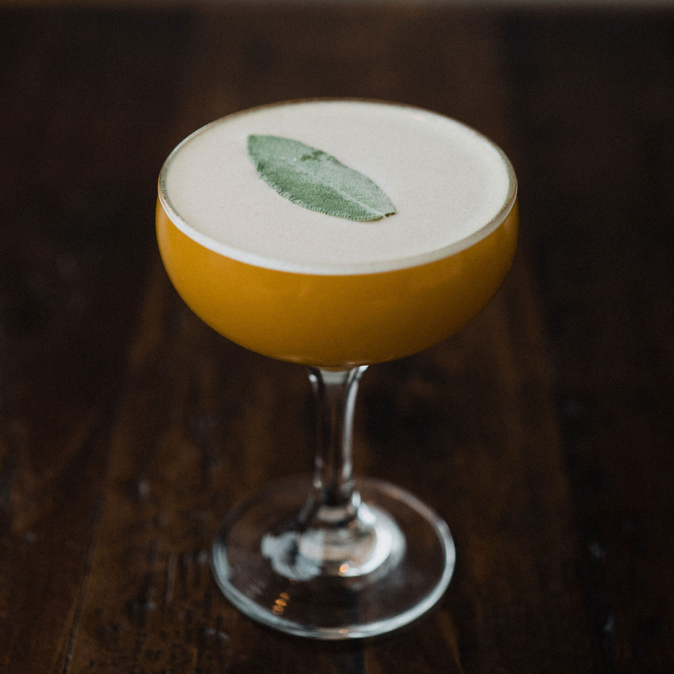 A golden cocktail with a frothy top and a single sage leaf garnish on a dark wooden table.
