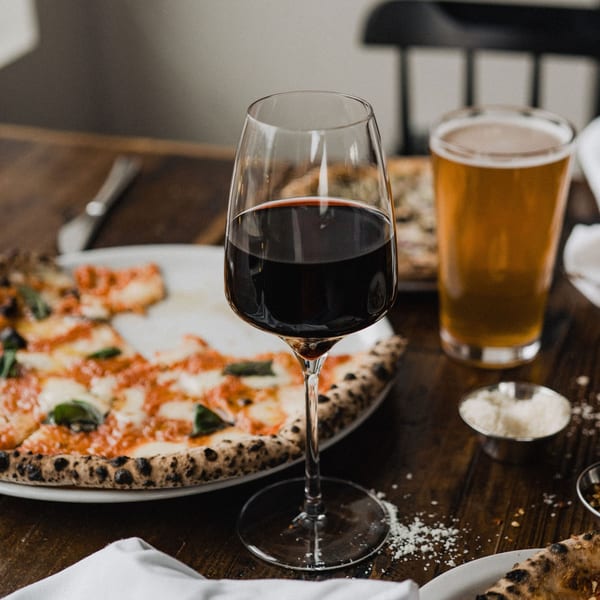 A glass of red wine and a beer next to a wood-fired pizza on a rustic wooden table.